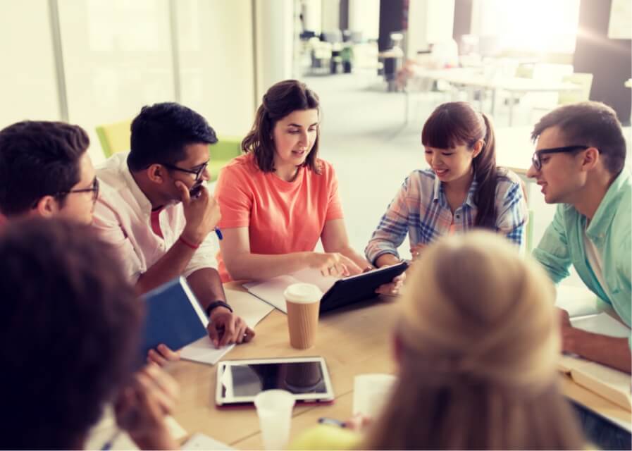 A group of people sit around a table looking at a tablet computer smiling and talking